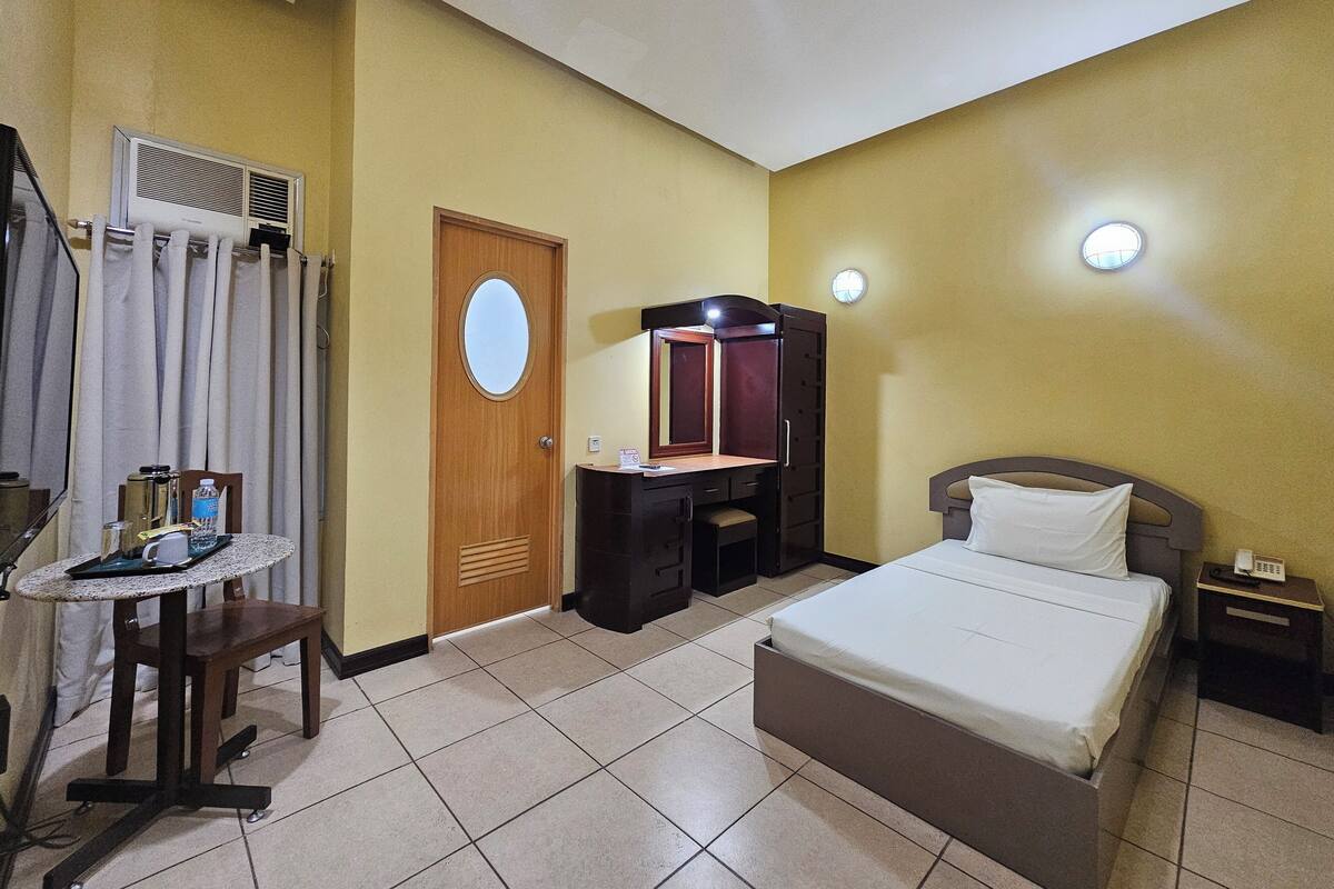 Habitación de hotel con una cama, un escritorio con un espejo, una silla, una mesita con una botella de agua y un teléfono en la mesita de noche.