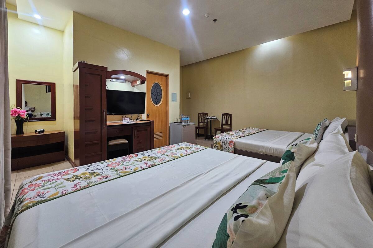 Habitación de hotel con dos camas, un escritorio con televisión de pantalla plana, un espejo, una pequeña mesa con sillas y una puerta con panel de vidrio decorativo.