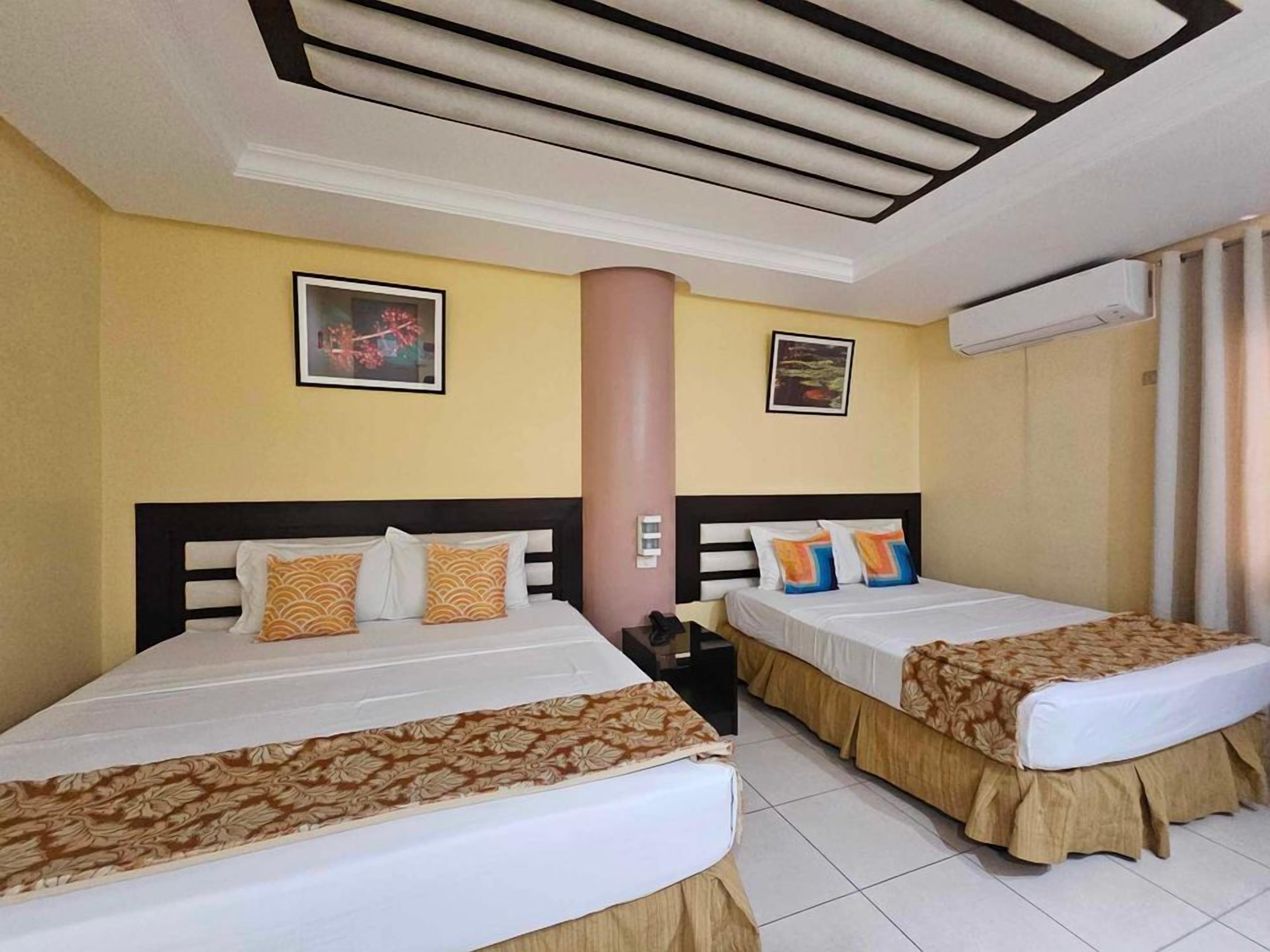 A hotel room with two beds, each with a patterned bedspread and pillows, a large pillar, and two framed pictures on the walls.