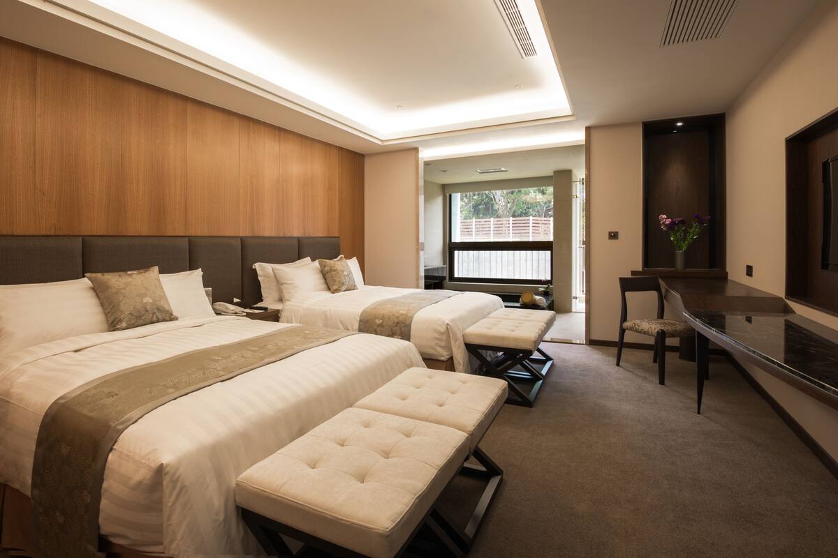 A modern hotel room with two beds, a desk, and a view of the outdoors.