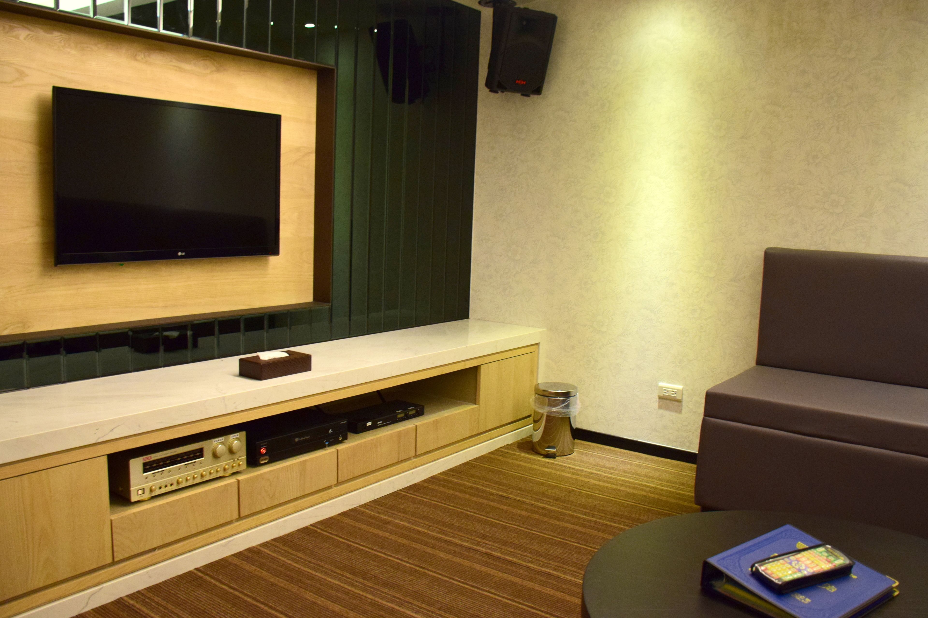 A modern living room with a flat-screen TV mounted on a wooden wall, a built-in entertainment unit, a brown leather sofa, and a small table with a book and remote control.