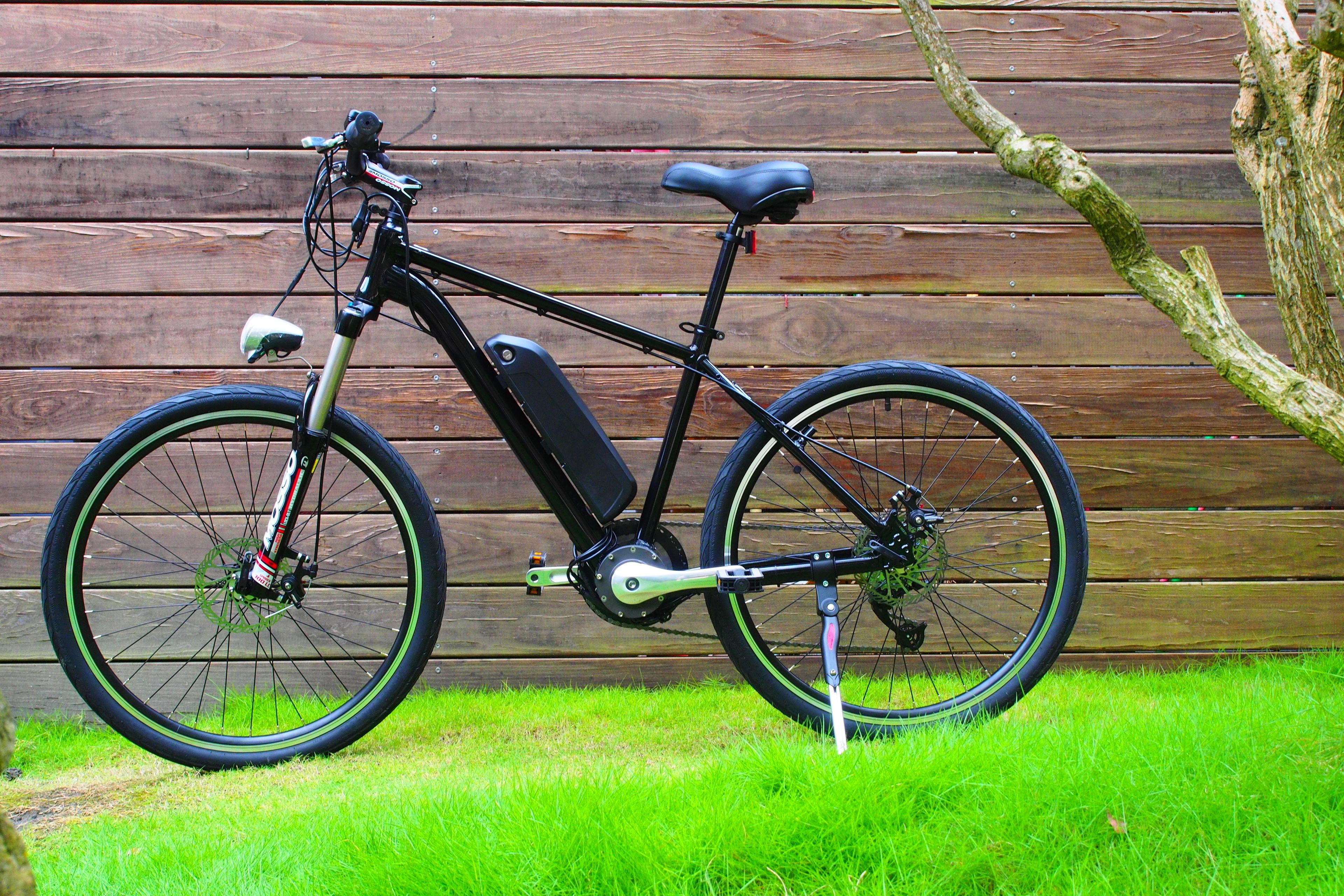 A stationary electric bicycle with a black frame, front and rear suspension, and a battery pack mounted on the down tube.