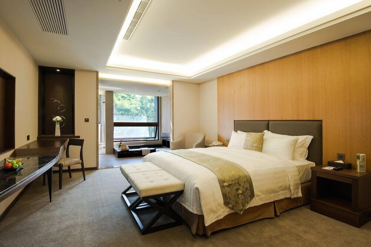 A modern hotel room with a large bed, a desk, and a view of greenery outside.