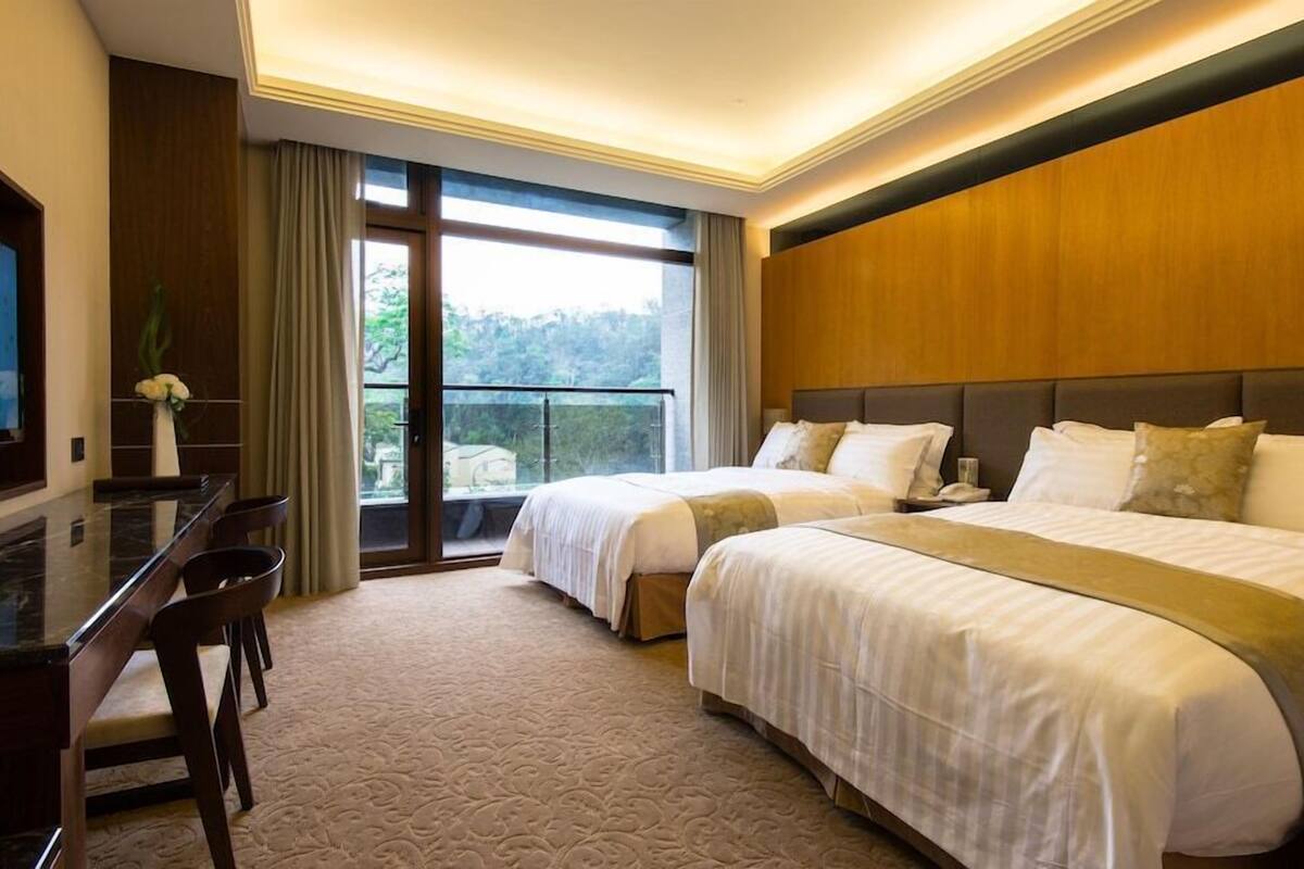 A hotel room with two beds, a desk, a chair, and a large window with a view.