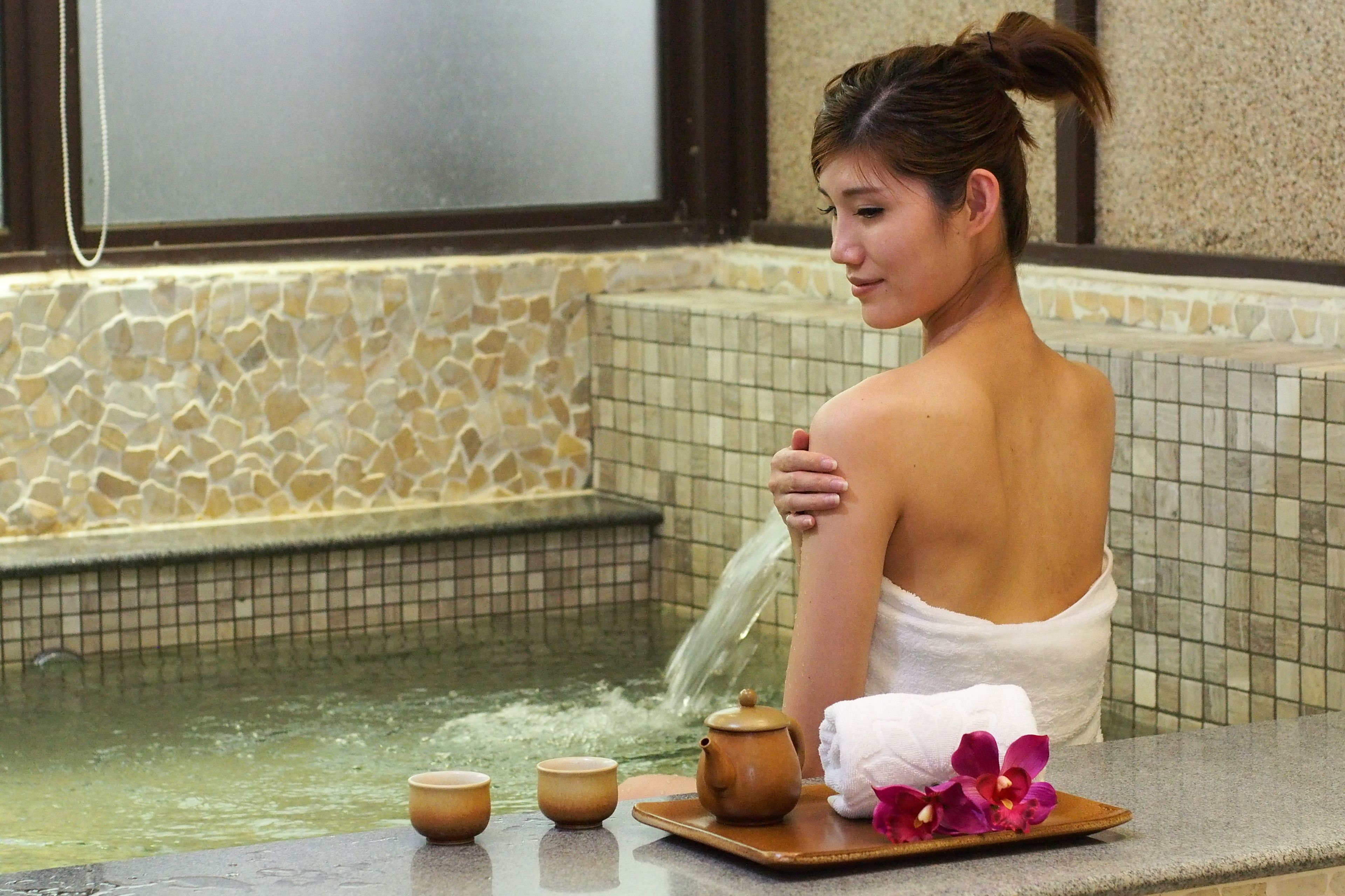 A person in a spa setting with a stone-tiled wall, a small pool, and a tray with a teapot and cups.