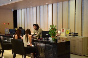 Una mesa de recepción con una recepcionista, tres huéspedes sentados y una planta en maceta.