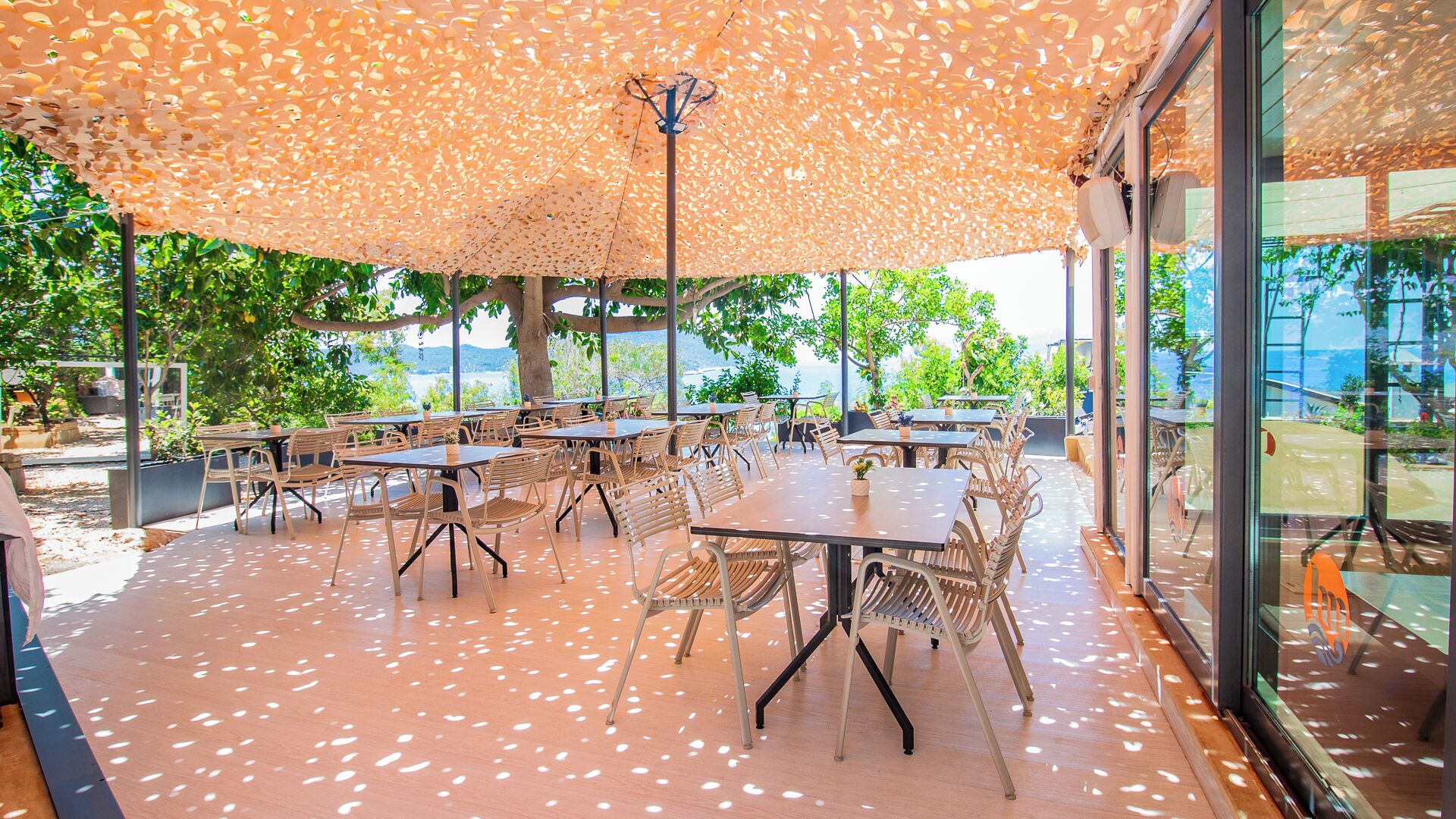 An outdoor dining area with tables and chairs, a pergola with a leaf-patterned canopy, and a view of the sea.