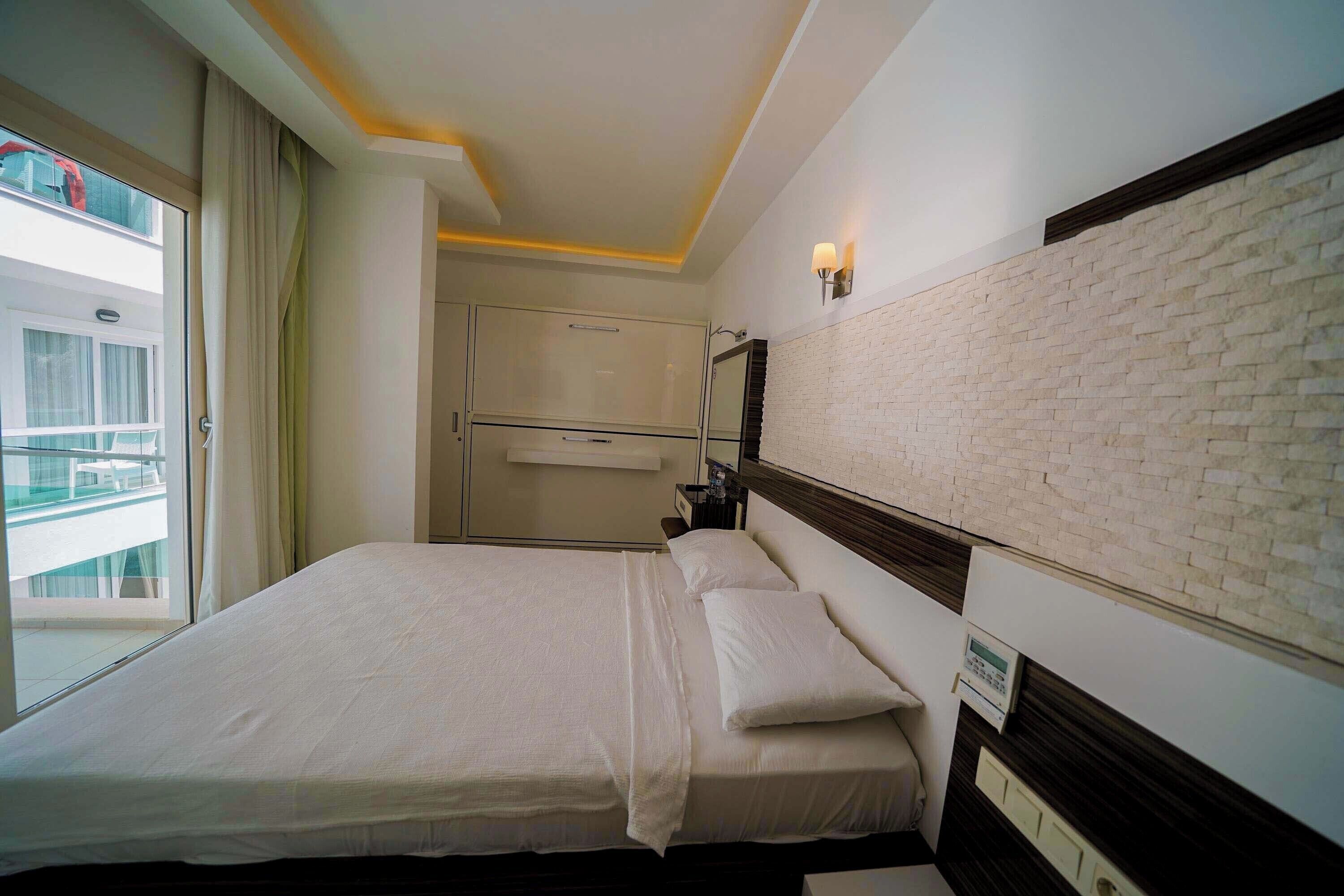 A hotel room with a bed, a nightstand, a wall-mounted light, and a view of the outside.