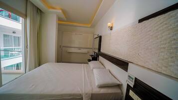 A hotel room with a bed, a nightstand, a wall-mounted light, and a view of the outside.