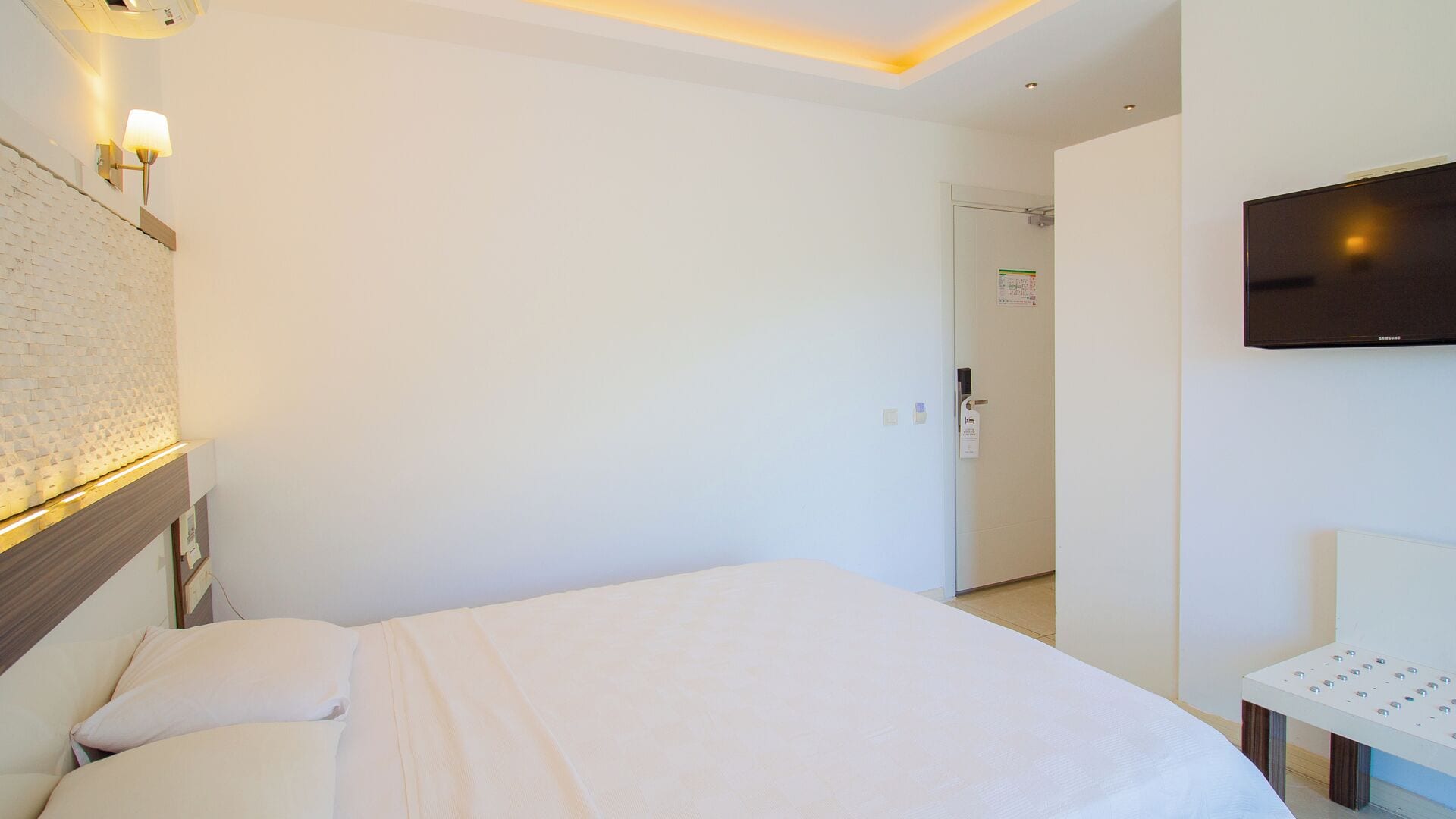 A hotel room with a bed, a desk, a flat-screen TV, and a wall-mounted lamp.