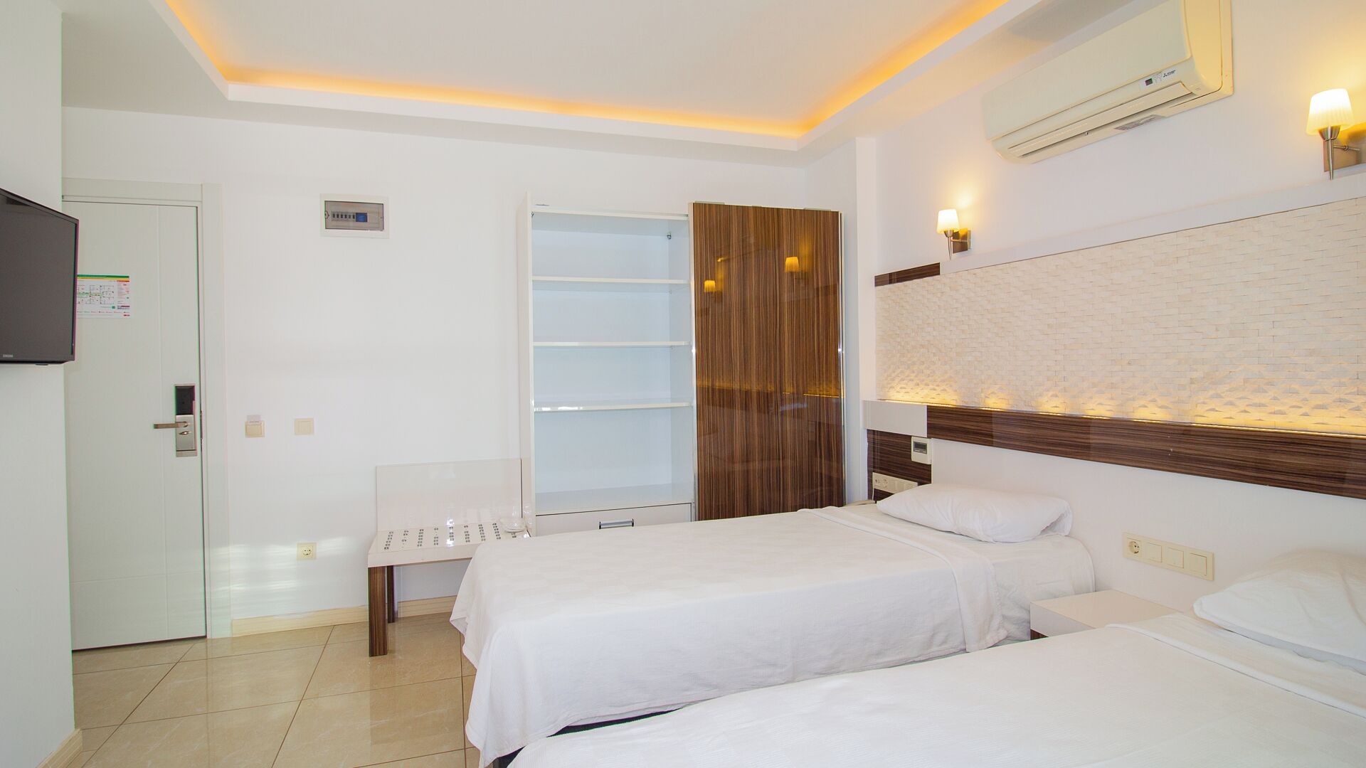 A hotel room with two beds, a wooden wardrobe, a glass cabinet, and a wall-mounted TV.