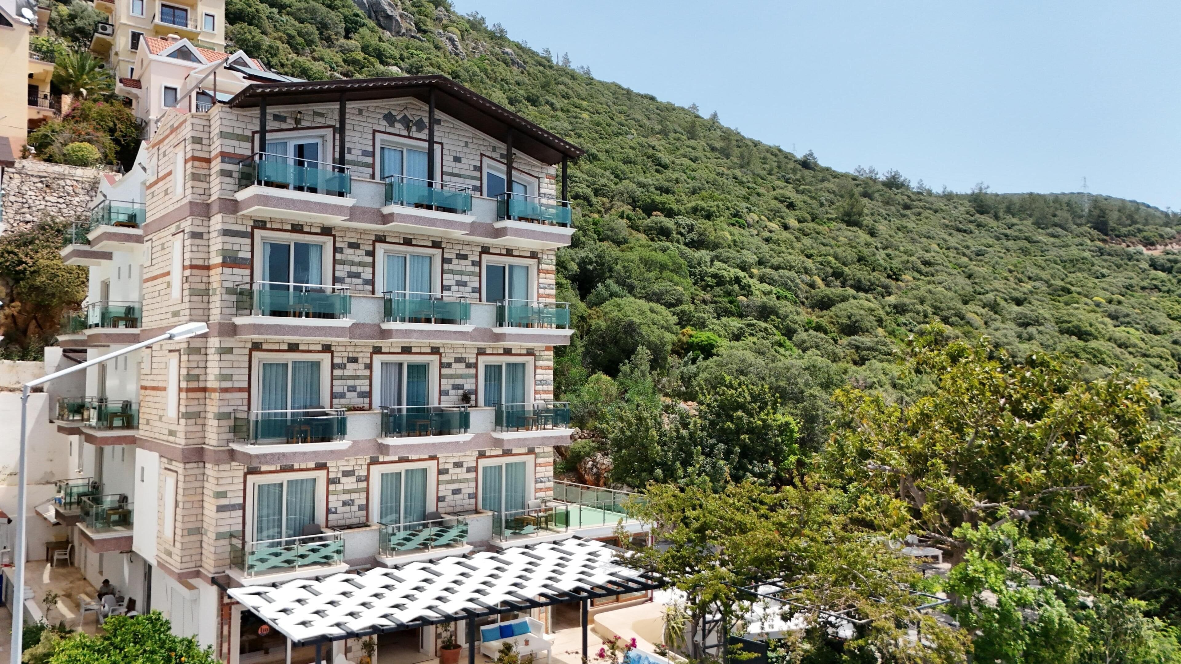 A multi-story hotel with balconies, a pool area, and a view of a forested hillside.