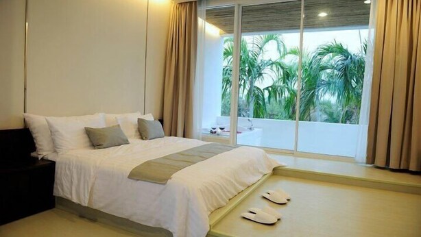 A modern hotel room with a large bed, a small bedside table, and a view of palm trees through a large window. - Ohana Resort and Restaurant (Rayong)