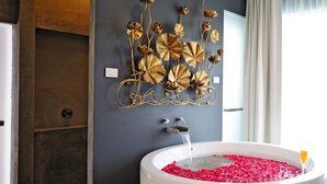 A modern bathroom with a large bathtub filled with rose petals, a wall-mounted gold floral art piece, and a window with curtains. - Ohana Resort and Restaurant (Rayong)