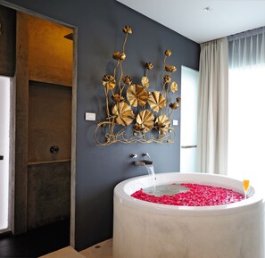 Et moderne badeværelse med et stort badekar fyldt med rosenblade, et guldmaleri med blomstermotiver på væggen og et vindue med gardiner.
