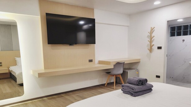 Et moderne hotelværelse med et fladskærms-tv monteret på en trævæg, et skrivebord med stol, en seng med et pænt foldet håndklæde og et badeværelse med marmorvæg.