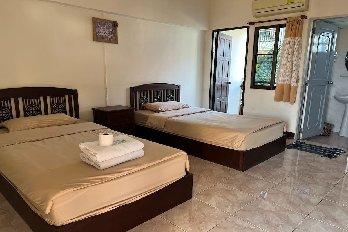 A hotel room with two beds, a small bedside table, a wall-mounted air conditioner, a window with curtains, and a bathroom visible through an open door.