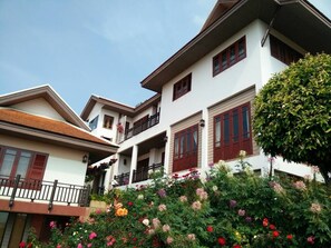A multi-story building with balconies, a garden with various flowers, and a clear sky. - PhuMorkDao Resort (Mae Rim)