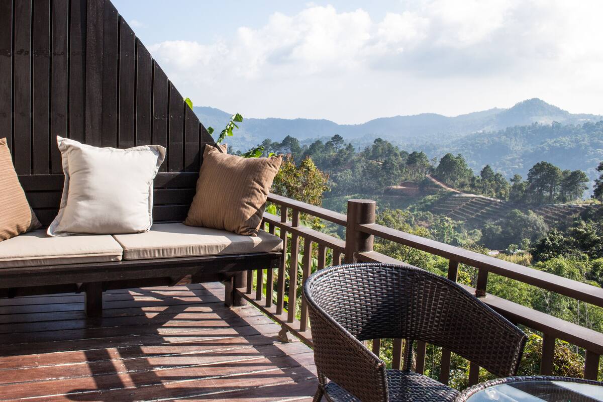 A wooden deck with a sofa and wicker chair, overlooking a mountainous landscape.