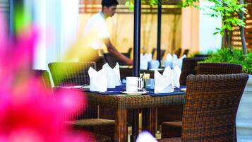 A dining area with wicker chairs and tables, each set with white napkins, a white mug, and a spoon.