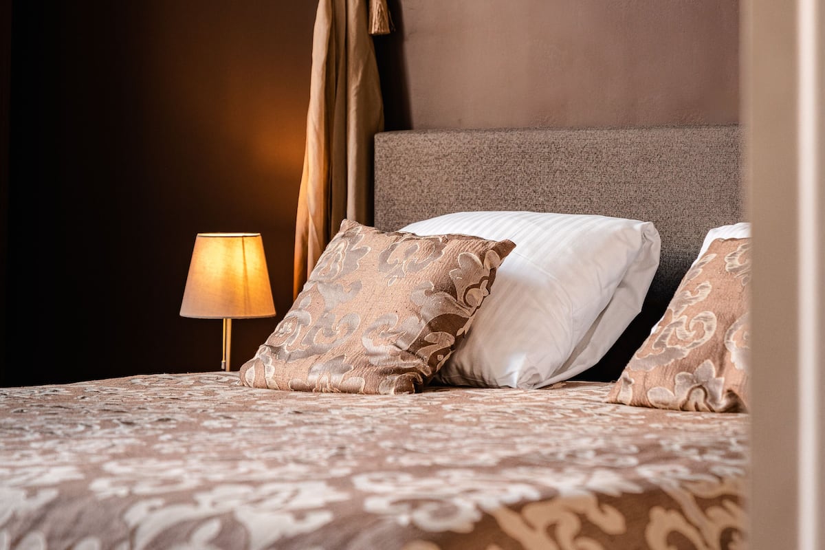 Una cama bien tendida con ropa de cama estampada, una lámpara de noche y un retrato en la pared.
