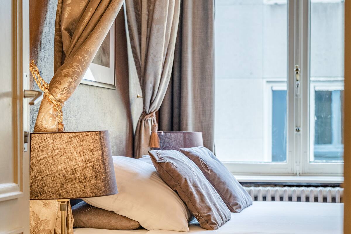Una cama bien hecha con edredón blanco, una mesita de noche de madera con una lámpara y vistas a un edificio a través de una ventana con cortinas.