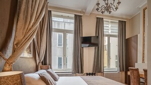 A hotel room with a large bed, a TV mounted on the wall, a chandelier, and a red carpet. - Hotel Van Eyck (Bruges)