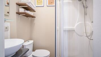 A modern bathroom with a white sink, toilet, and shower. There are towels on a shelf and framed artwork on the wall.