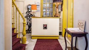 A staircase with yellow railings, a red patterned carpet, a white reception desk, and a statue. - Hotel Van Eyck (Bruges)