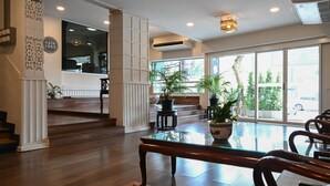 A spacious interior with a wooden floor, a glass-top table, and several chairs. There are potted plants and a large window providing a view outside.