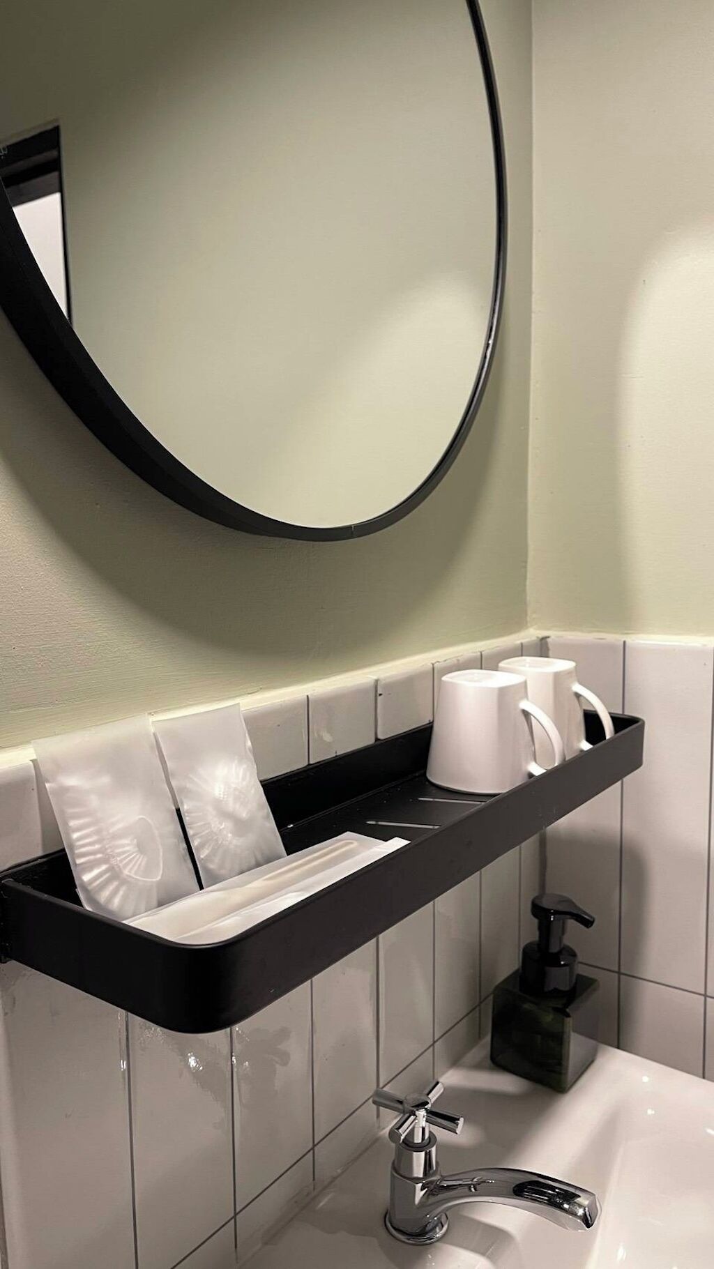 A modern bathroom with a black countertop, white tiles, a round mirror, and a black soap dispenser.