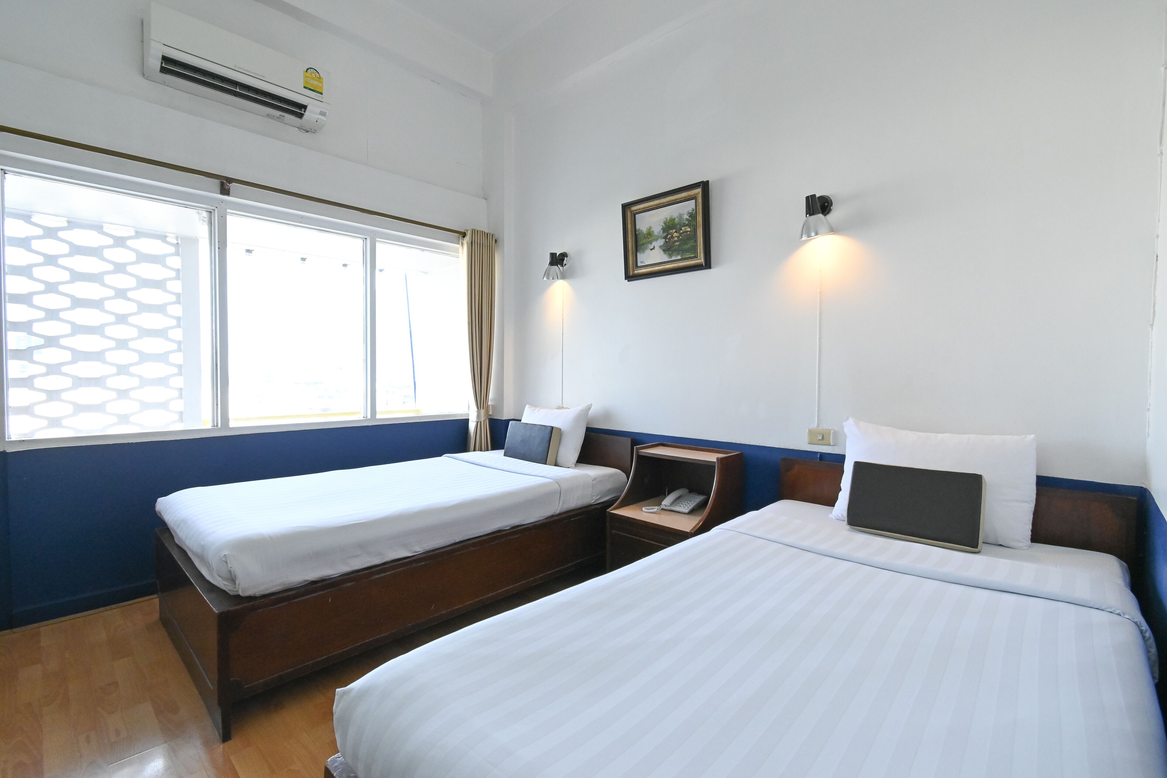Una habitación de hotel con dos camas, una ventana con persiana estampada, una mesita de noche con lámpara y un cuadro colgado en la pared.