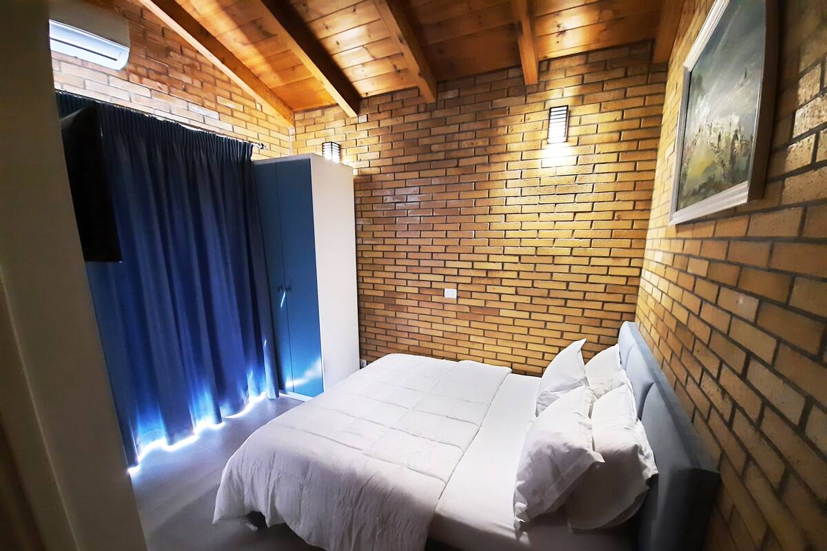 A bedroom with a brick wall, a bed with white bedding, a blue wardrobe, and a painting on the wall.