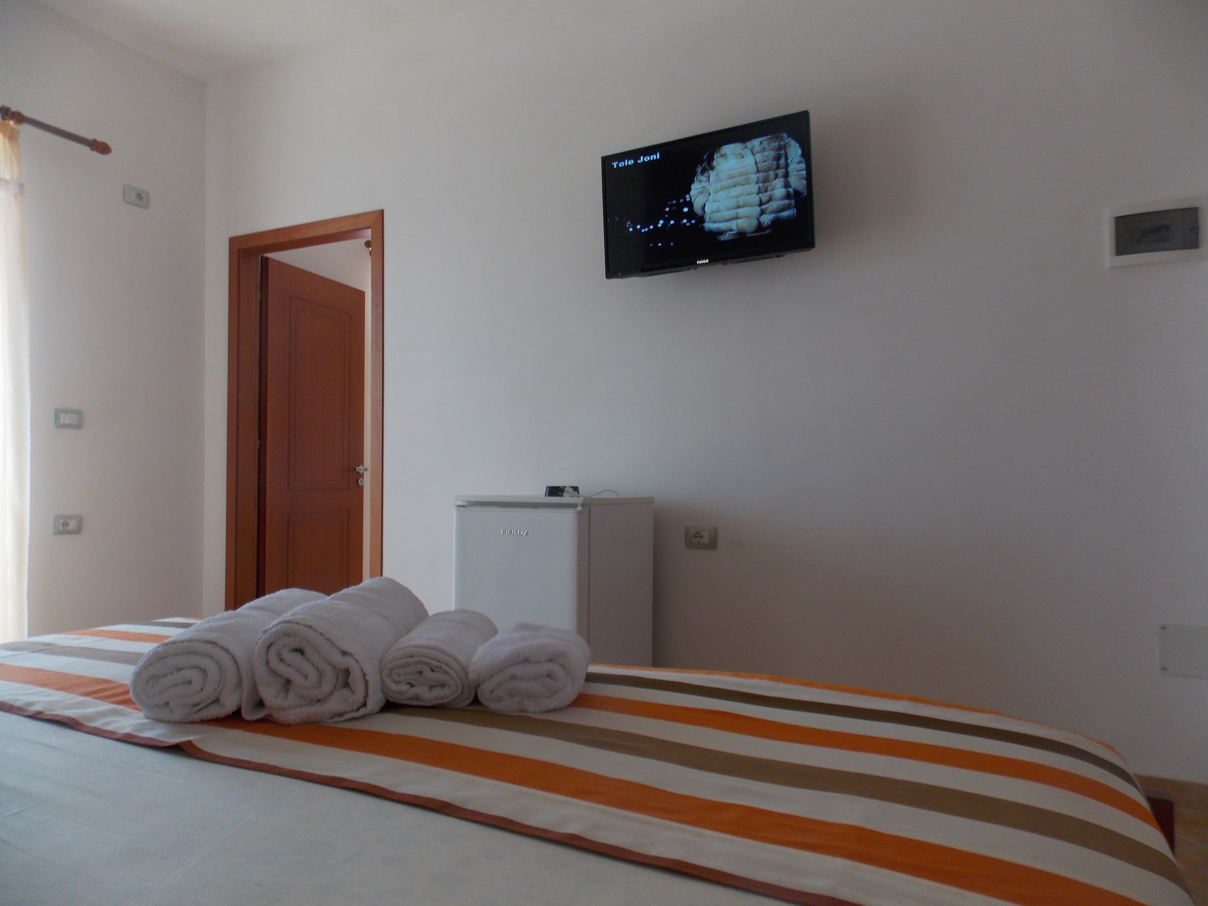 Una habitación de hotel con una cama, un televisor montado en la pared, una puerta y un pequeño electrodoméstico blanco.