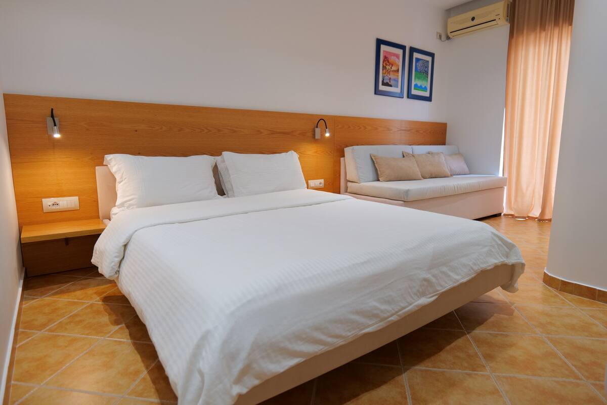 A modern bedroom with a large bed, a wooden headboard, two bedside lamps, a small sofa, and a wall-mounted air conditioner.