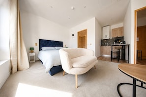 A bedroom with a bed, a chair, a small table, a window with curtains, and a kitchenette in the background. - HDG Appart's Hotel Gare de L'Isle Adam (Parmain)