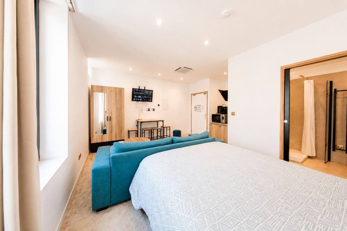 A modern bedroom with a large bed, a seating area with blue sofas, a desk, a TV, and a shower area.