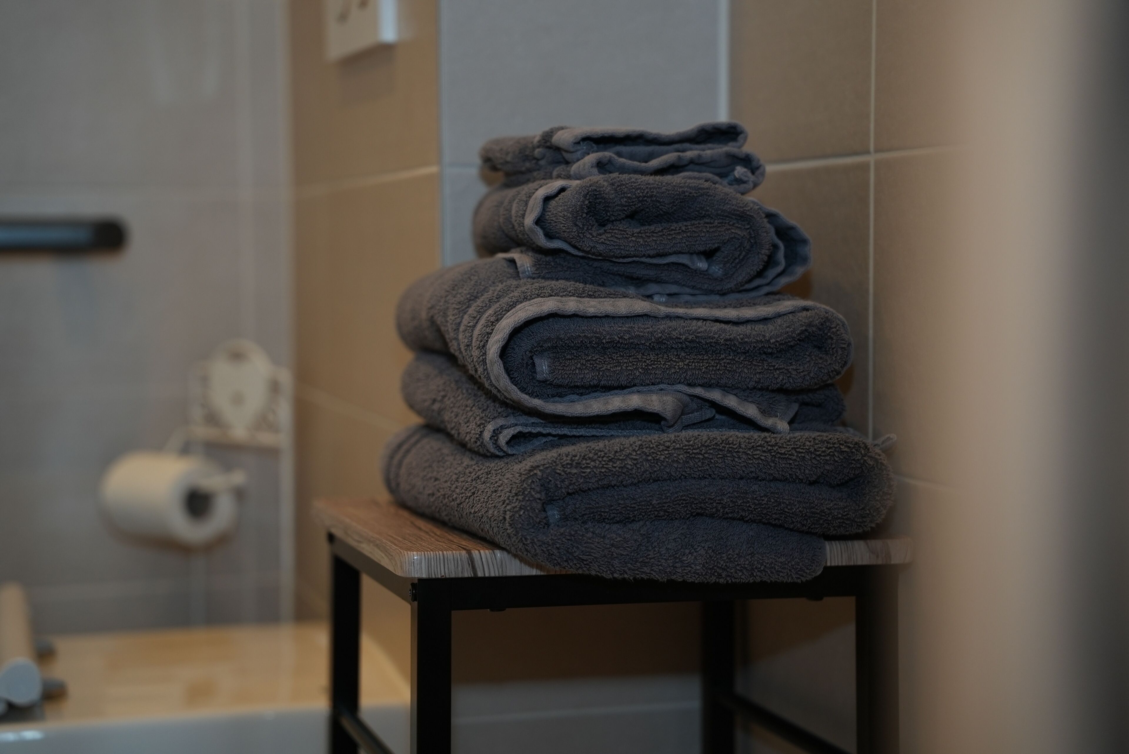 Une pile de serviettes pliées est posée sur une table en bois dans une salle de bain, avec un rouleau de papier toilette en arrière-plan.