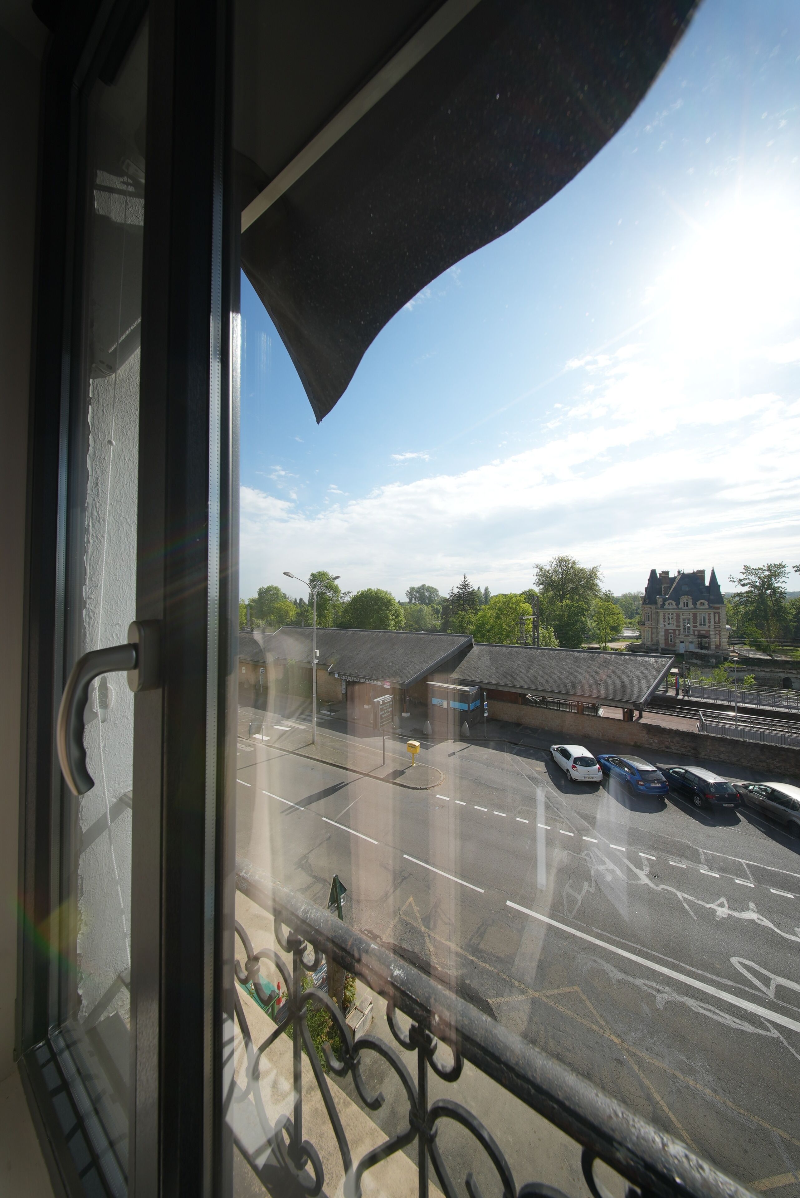 Blick aus einem Fenster mit Metallgeländer, ein Parkplatz und ein Gebäude mit geneigtem Dach im Hintergrund.