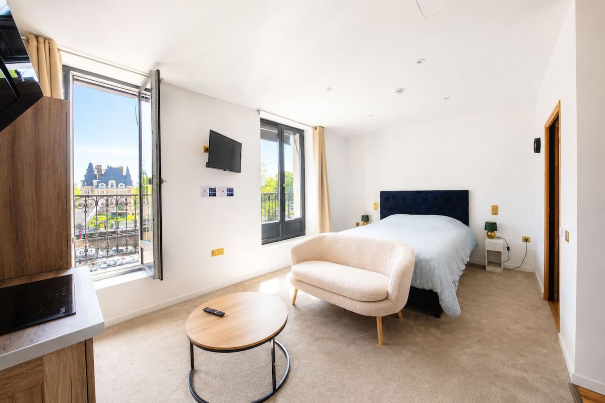 A modern hotel room with a bed, a TV, a small table, and a view of a building through large windows.