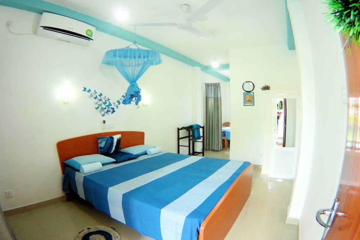 A bedroom with a bed, a ceiling fan, an air conditioning unit, a small table, and a chair.