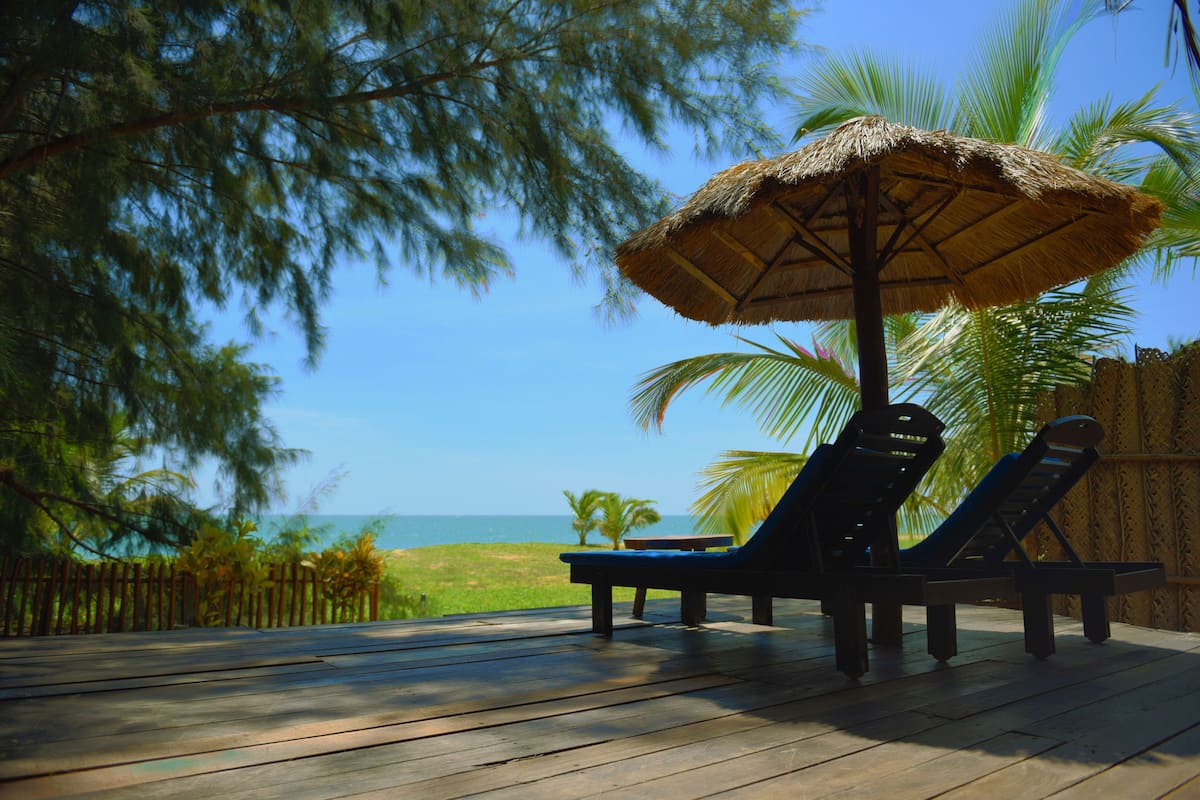 A wooden deck with two lounge chairs and a thatched umbrella, overlooking a beach and palm trees.