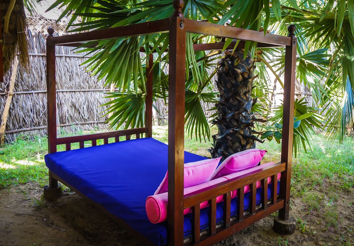 A wooden bed with a blue mattress and pink pillows, surrounded by lush green foliage.