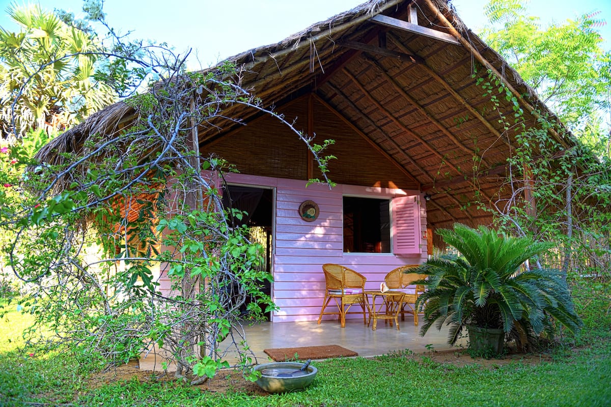 A rustic thatched-roof structure with a pink exterior, a porch with wicker chairs, and a small garden area.