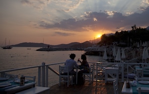 A sunset view from a deck with tables and chairs, overlooking a marina with boats and a distant mountain range.