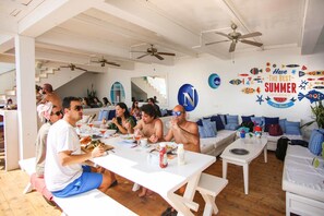 A dining area with white tables and benches, ceiling fans, and a wall with a summer-themed mural.