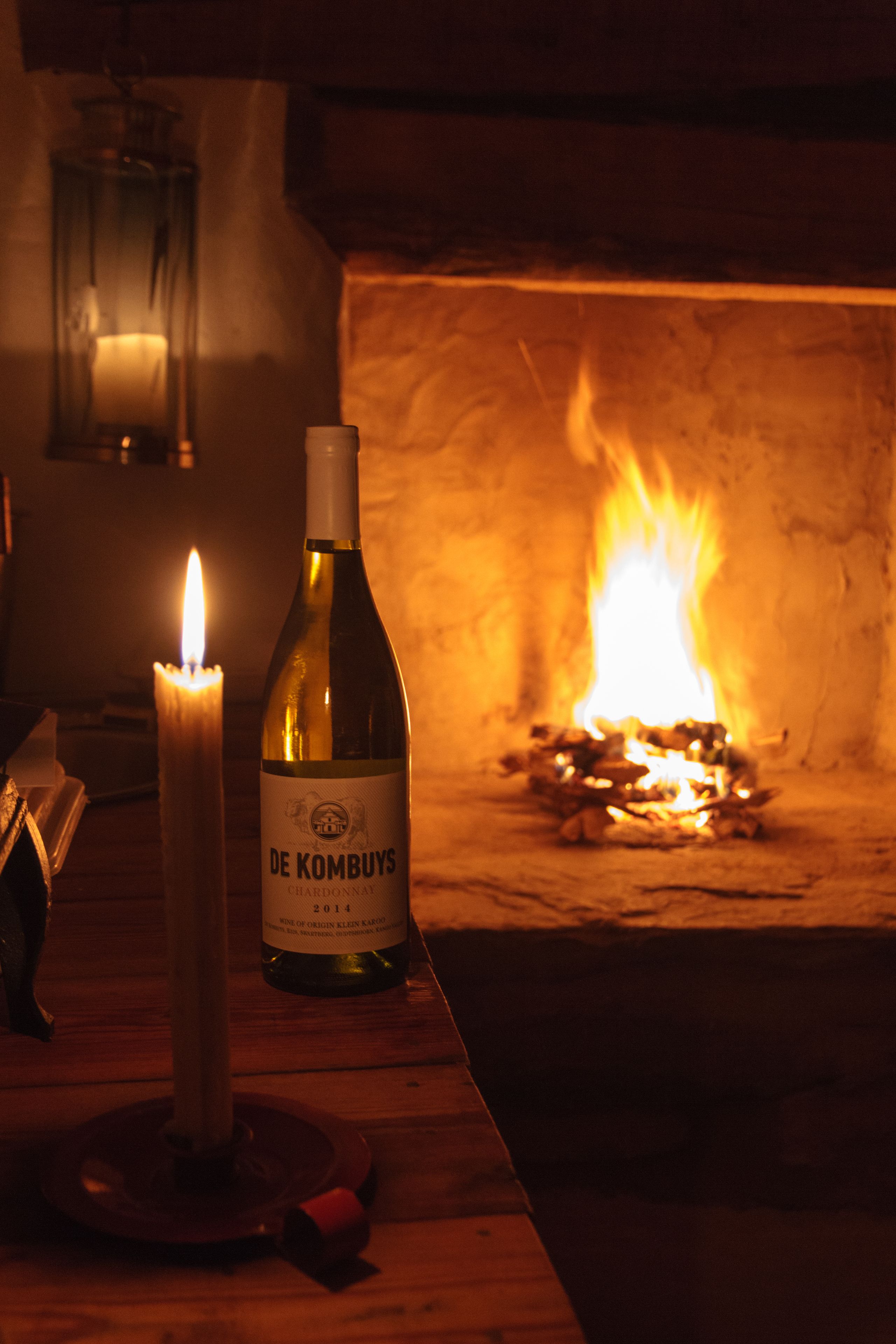 A bottle of De Krombys wine and a lit candle on a wooden table in front of a fireplace.