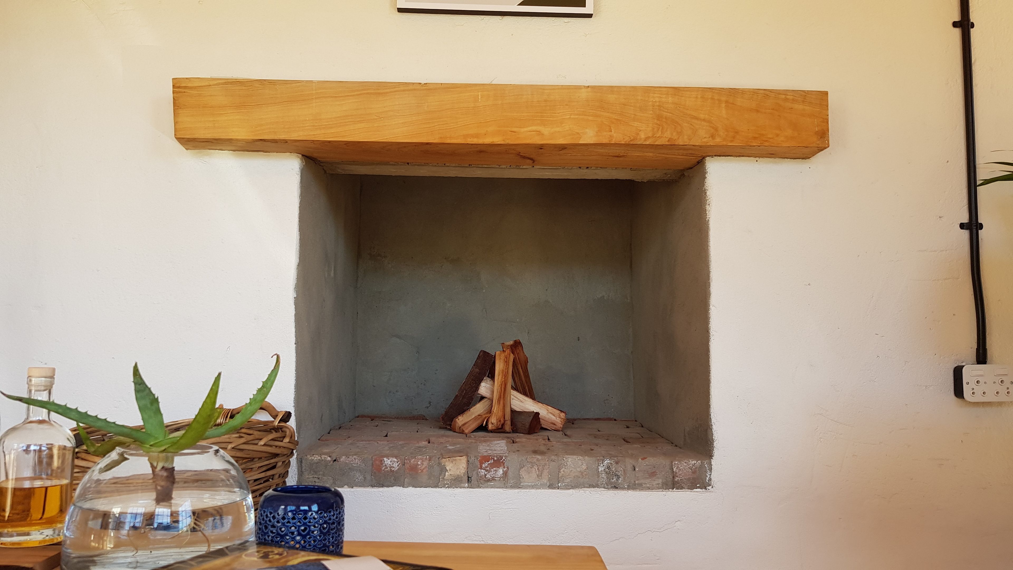 Une cheminée avec un manteau en bois, un foyer en briques et des bûches à l’intérieur. Deux vases avec des plantes sont posés sur une table devant la cheminée.