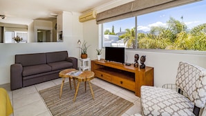 A modern living room with a sofa, a television, a wooden entertainment unit, and a large window with a view of greenery. -  Morgen Guest House (Cape Town)