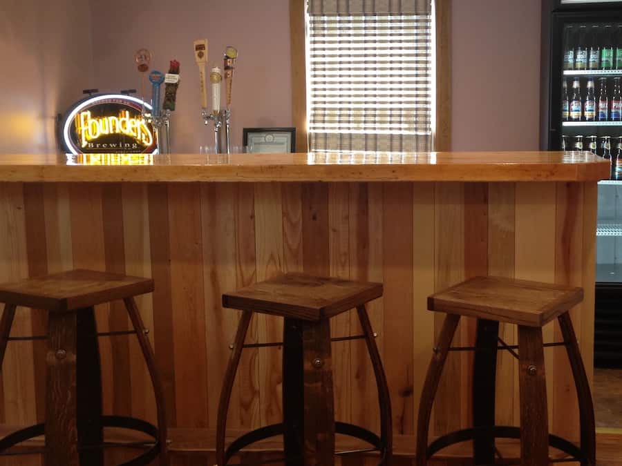 A bar area with wooden stools, a neon sign, and a refrigerator stocked with beverages.